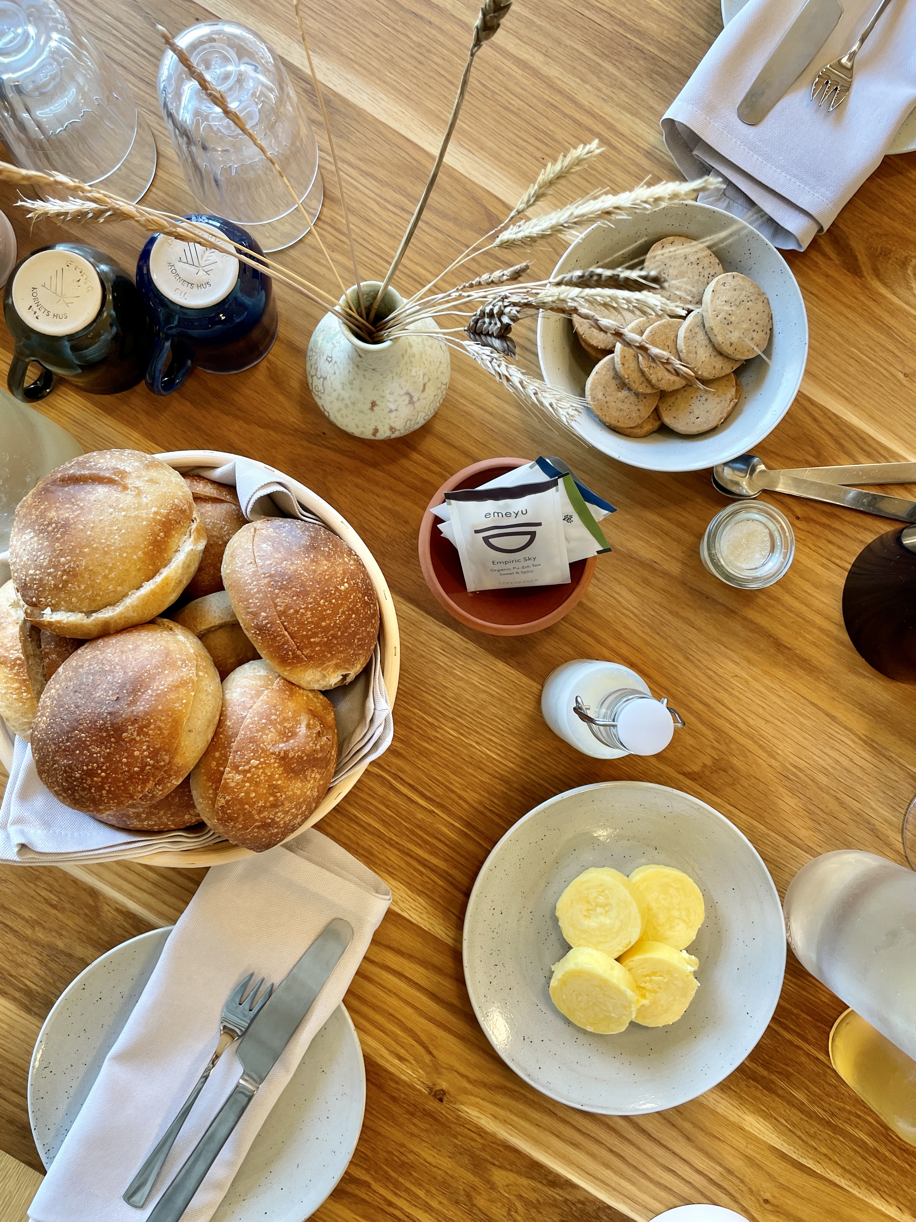bord dækket med tallerkener, kopper og glas samt boller i brødkurv, smør på tallerken og småkager i skål