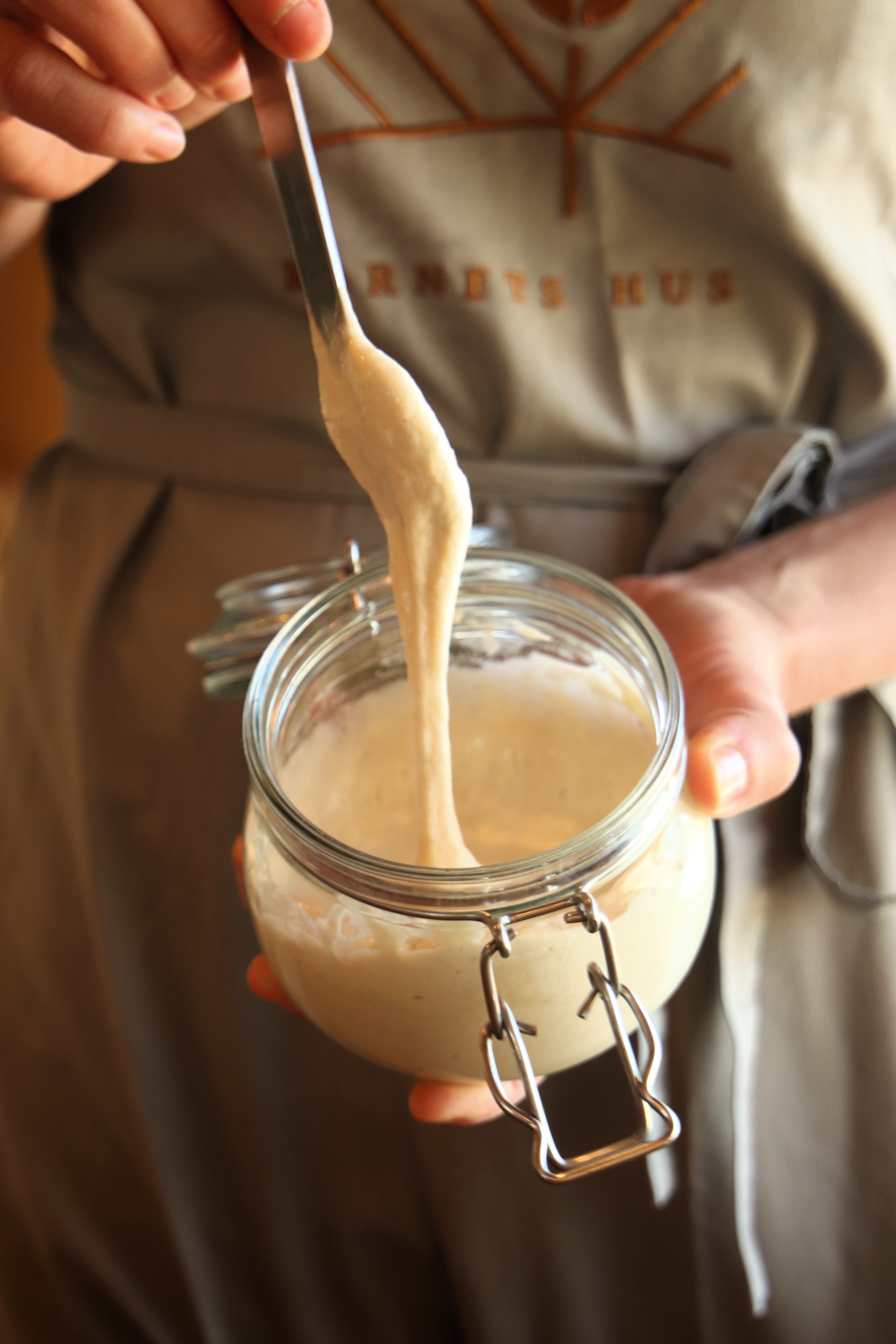 surdej i glas og hånd, der holder en ske med surdejen dryppende fra