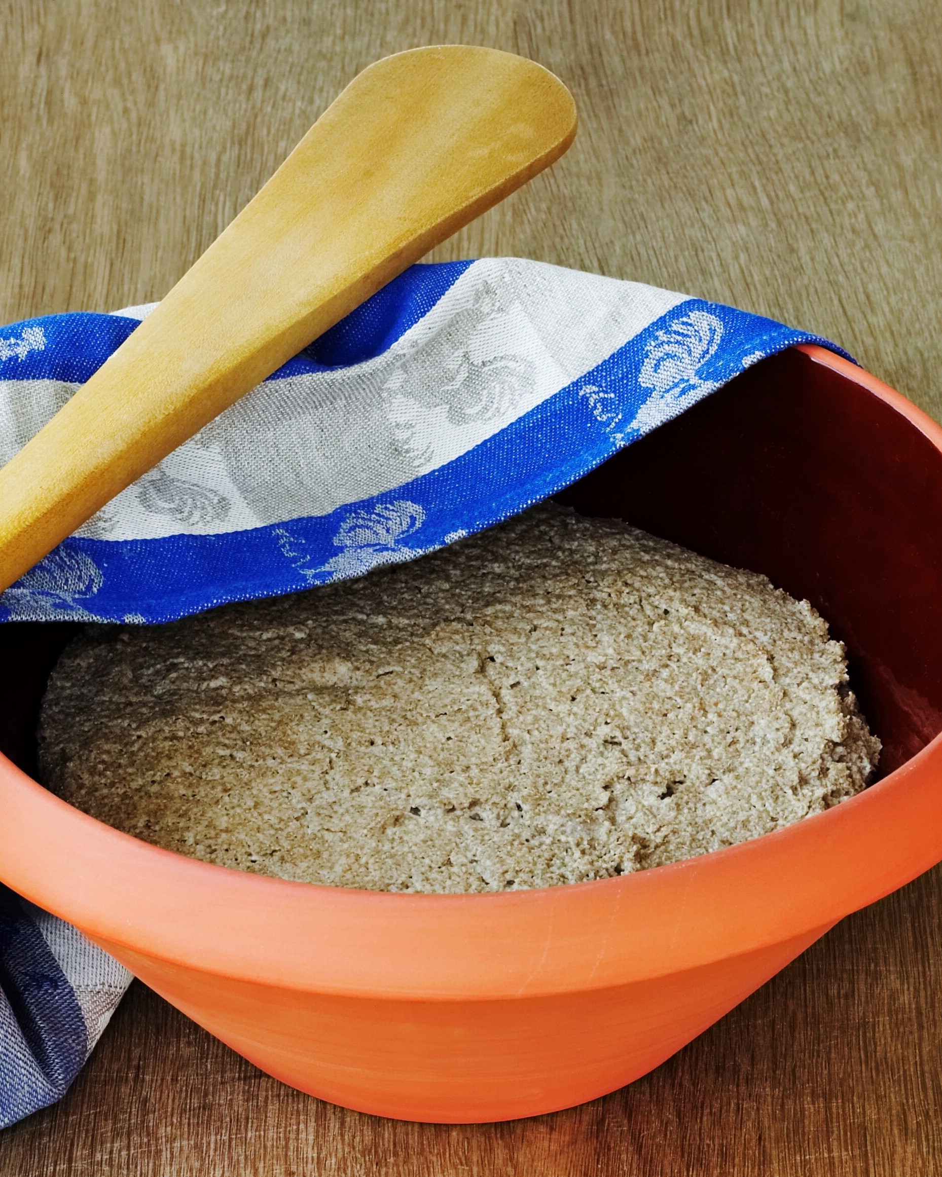 rugbrødsdej i lerfad med viskestykke trukket halvt over og røreske i træ hvilende ovenpå