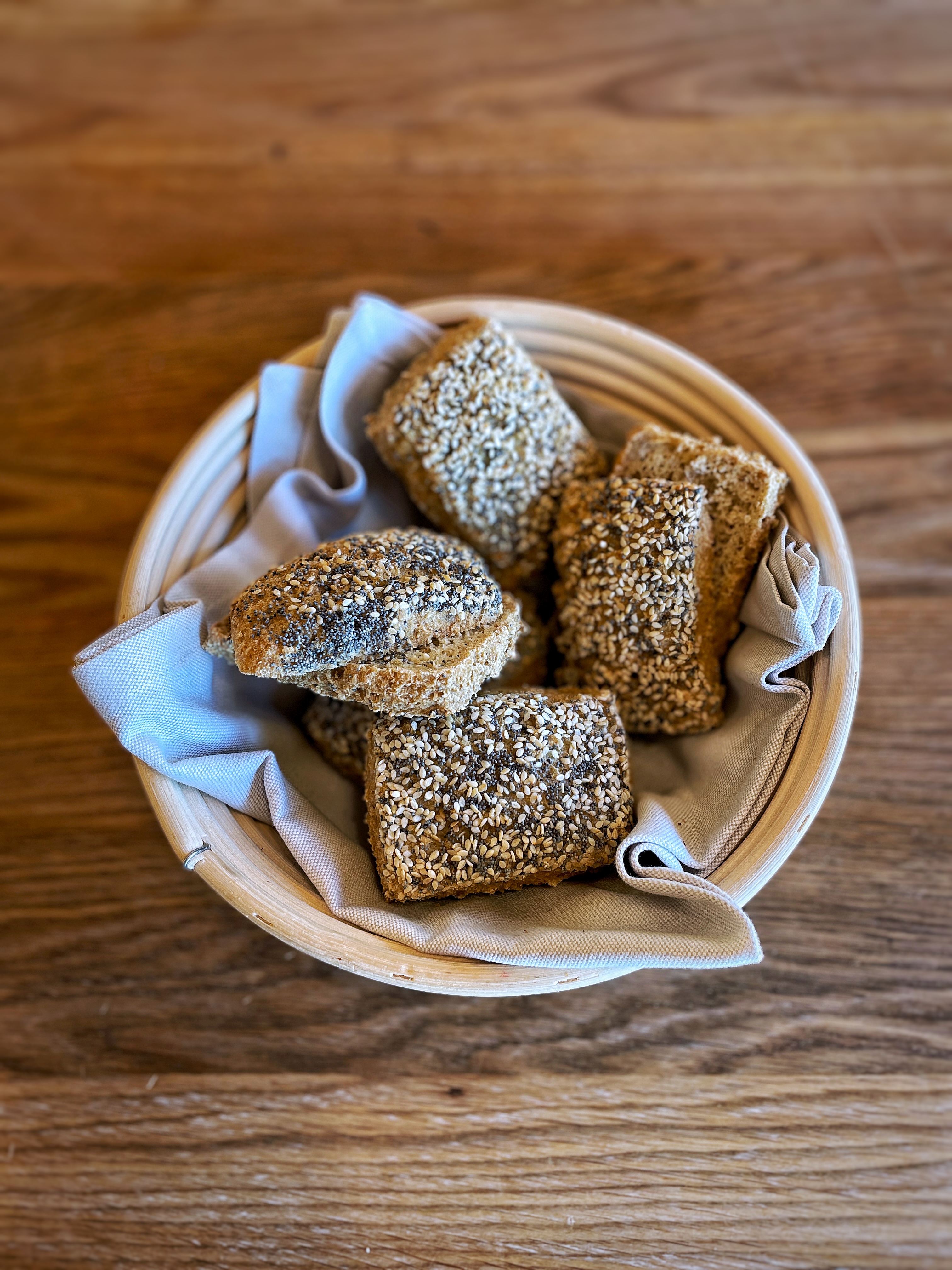 boller med drys af frø og kerner ligger i brødkurv med klæde på et træbord